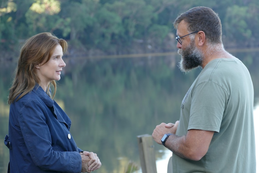 Rachel Griffiths is pictured side-on, speaking with a man who has his hands clasped. There is a river behind them. 