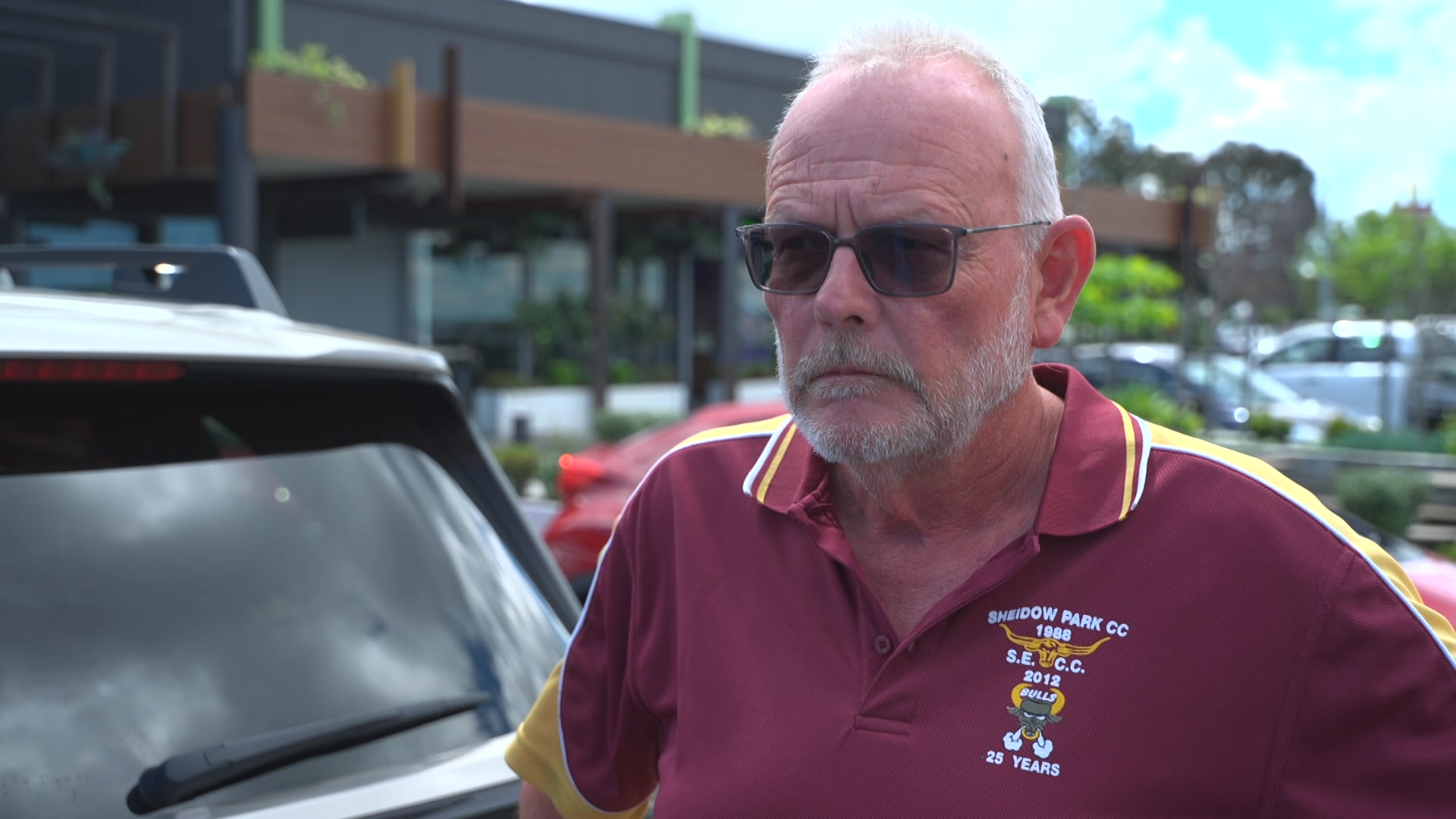 A man in a maroon tshirt with his hands on his hips has a neutral expression to the camera.