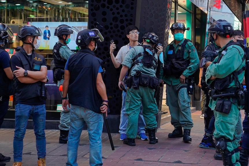 A young man holds a mobile phone in his right hand as he is searched while surrounded by heavily armed police.