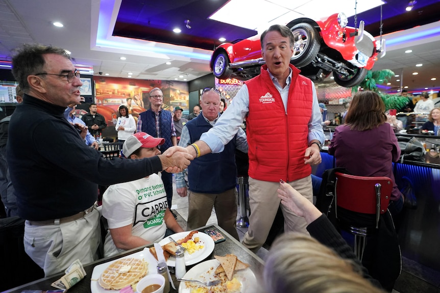 A middle-aged man in a red vest shakes hands with another man in a flashy, American-style diner.