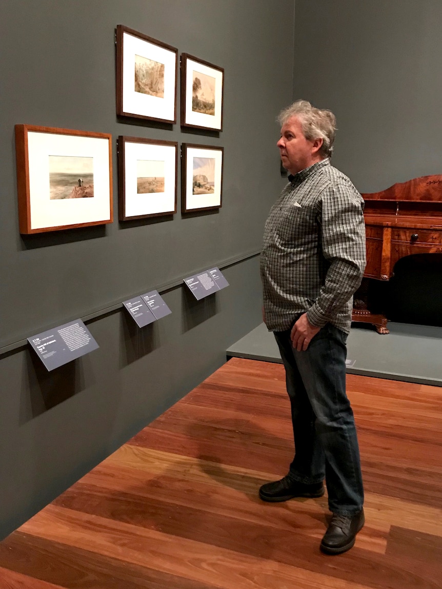 A man stands in an art gallery looking at pieces on the wall.