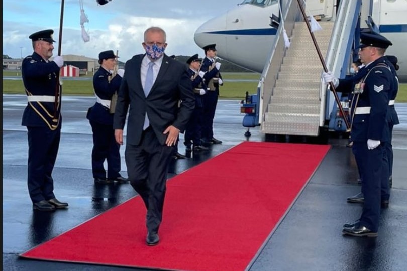 Australia Prime Minister Scott Morrison walks on a red carpet flanked by military personnel as he leaves a plane.