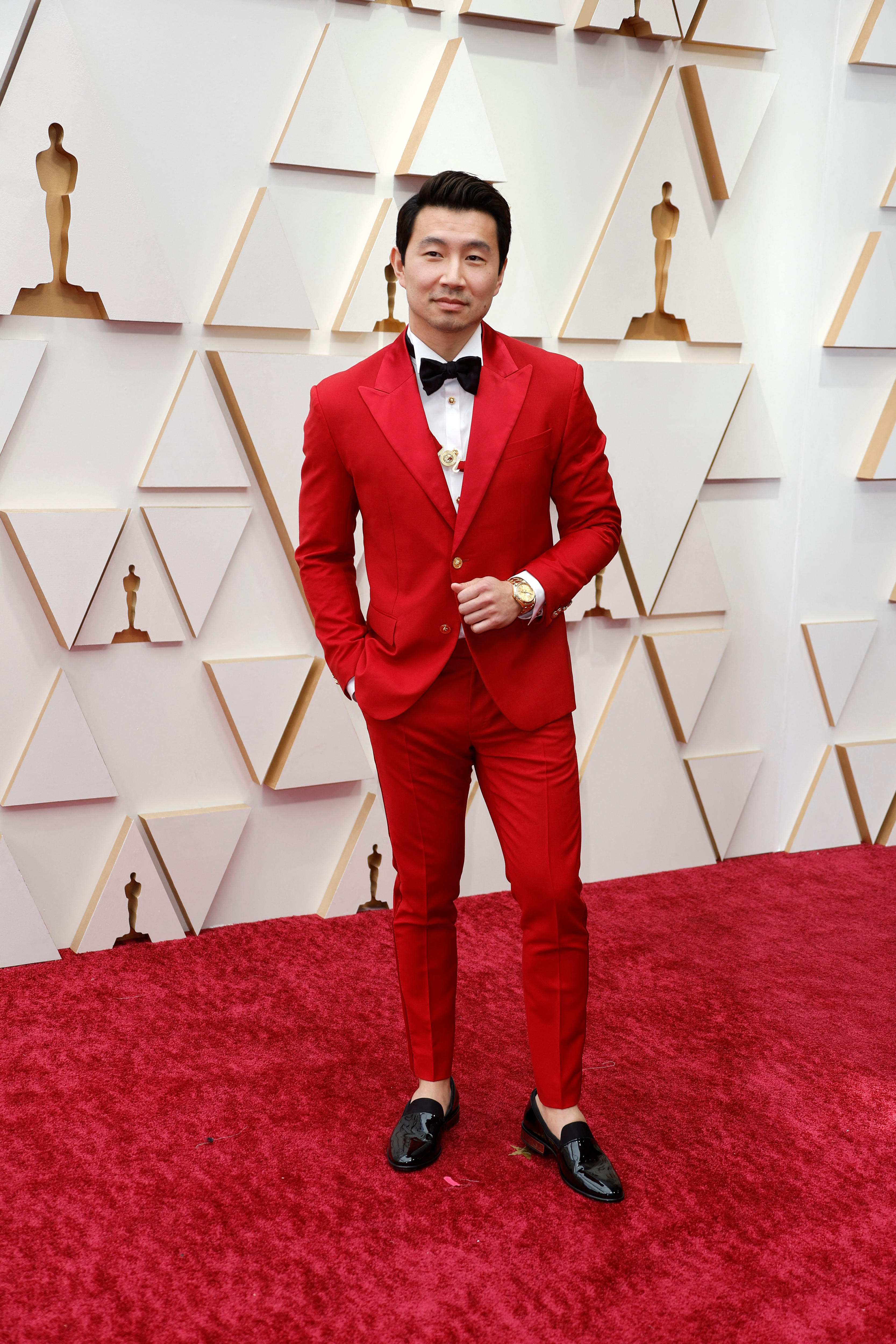 A man wearing a red suit and black loafers stands on a red carpet in front of a white media wall for the Oscars.