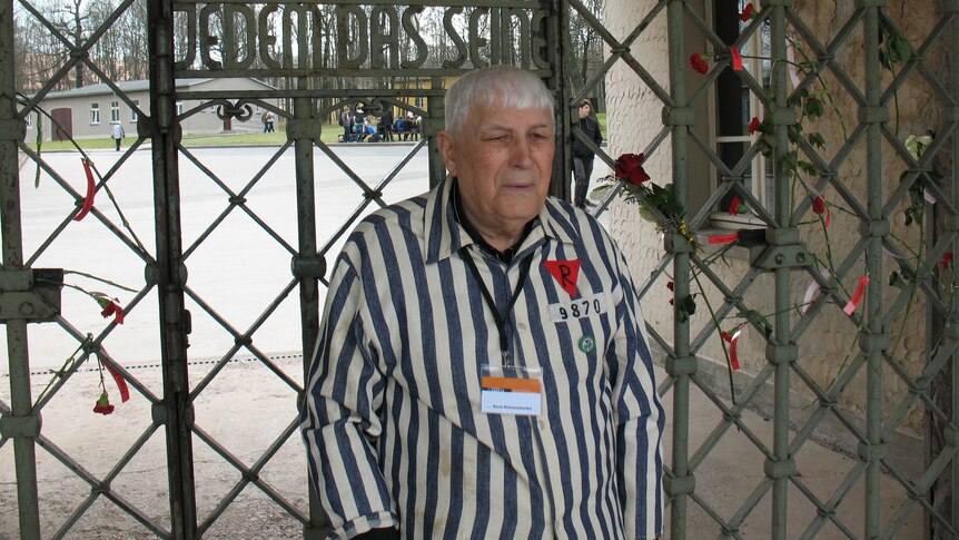 An old man in a vertically striped shirt poses for a photo before a metal gate.