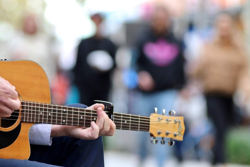 A busker plays an acoustic guitar as people observe while walking past.