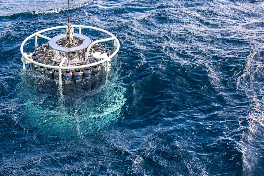 A circular scientific instrument with water bottles attached being lowered into the ocean.