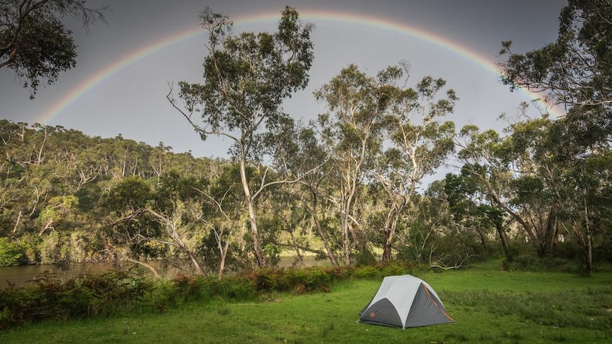 A small tent in a grassy field next to a river, with a rainbow and gum trees in the background.