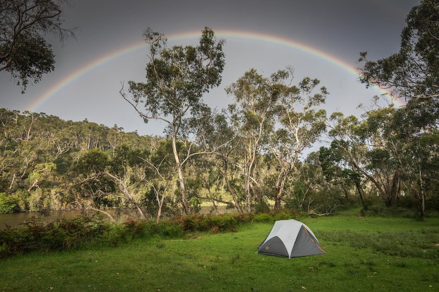 A small tent in a grassy field next to a river, with a rainbow and gum trees in the background.