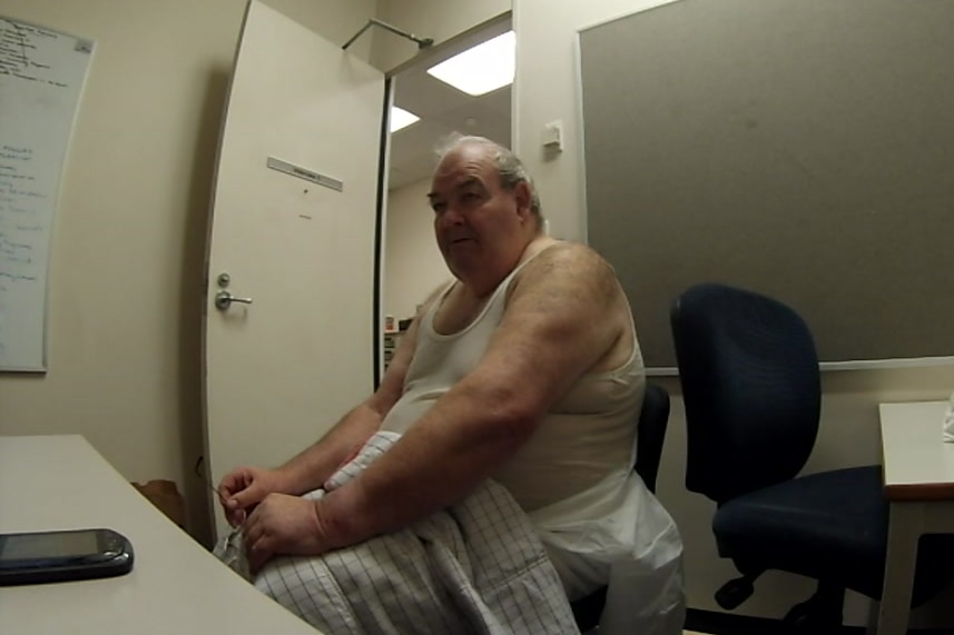 A man wearing a singlet with a blanket over his lap in an interview room.