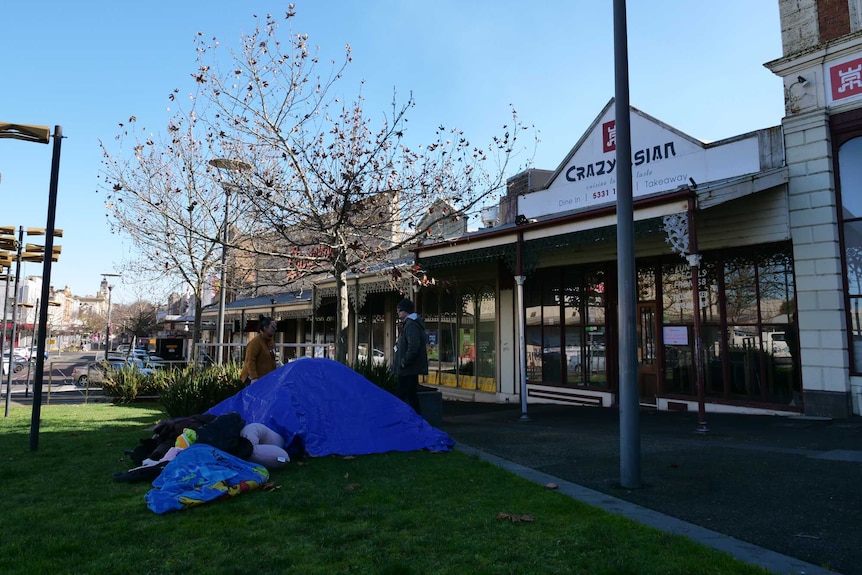 A tent and belongings on grass in front of some shops. 