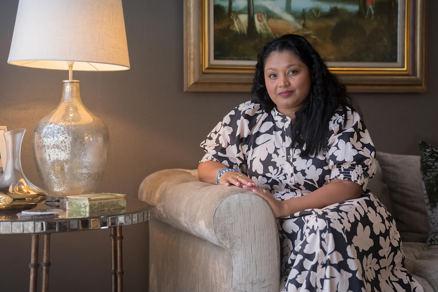 A woman with black hair sits on a chair in a lounge room setting