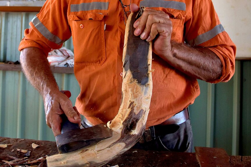 Roebourne Pastor Marshall Smith wearing a high-vis vest crafts a boomerang using an axe from a piece of local wood.