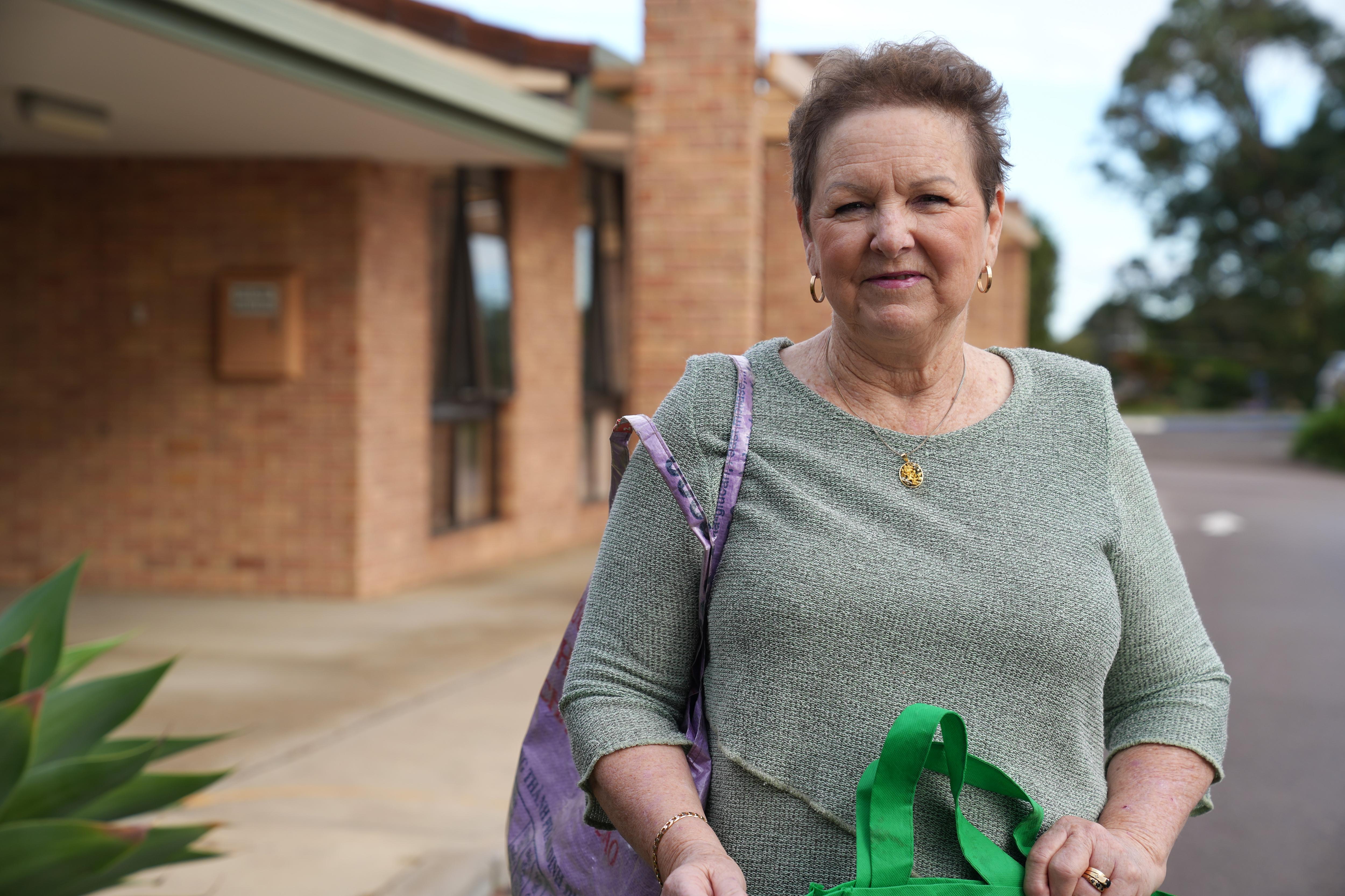 She holds two bags and stands by a brick building