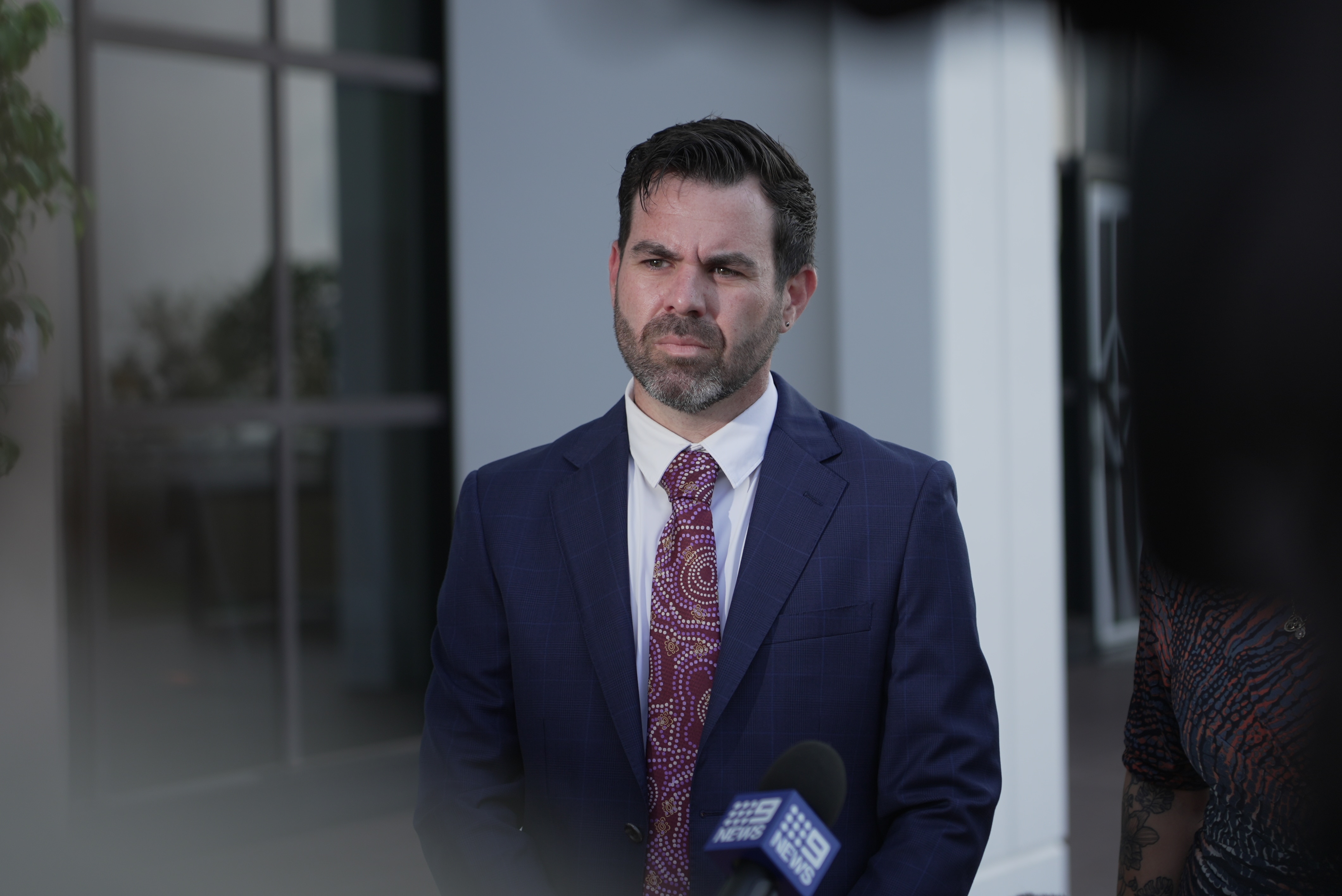 A man in a suit standing in front of a microphone, outside a building, and looking serious.