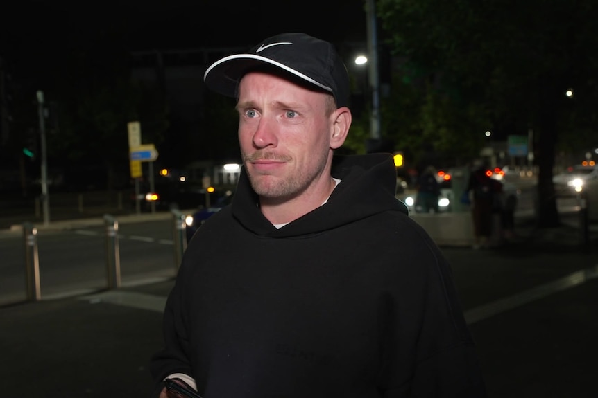 Un hombre que lleva una gorra de béisbol negra y una sudadera con capucha negra se para en una calle oscura en las afueras de Melbourne Park