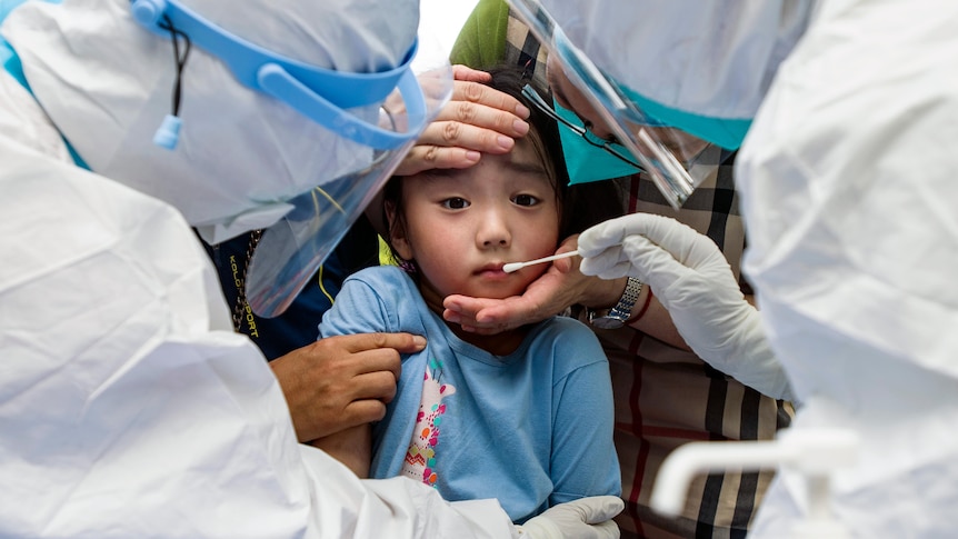 A child surrounded by three people in personal protective gear and goggles with a nose swab.