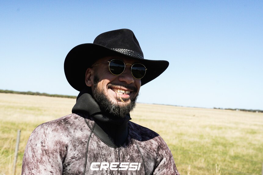 A man in a cowboy hat wearing a wetsuit chewing on a piece of straw smiling.