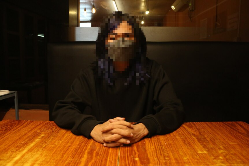 A young student, whose face is pixelated to avoid identification, sits at a table with hands folded in front of them.