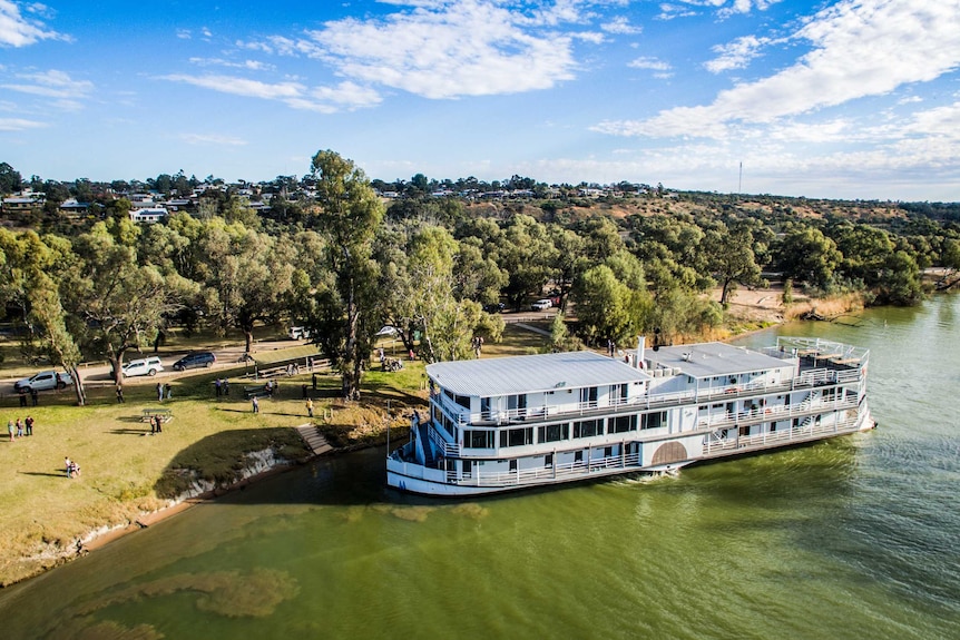 An aerial shot of a grand old paddle-steamer moored in front of parkland at the edge of a river.