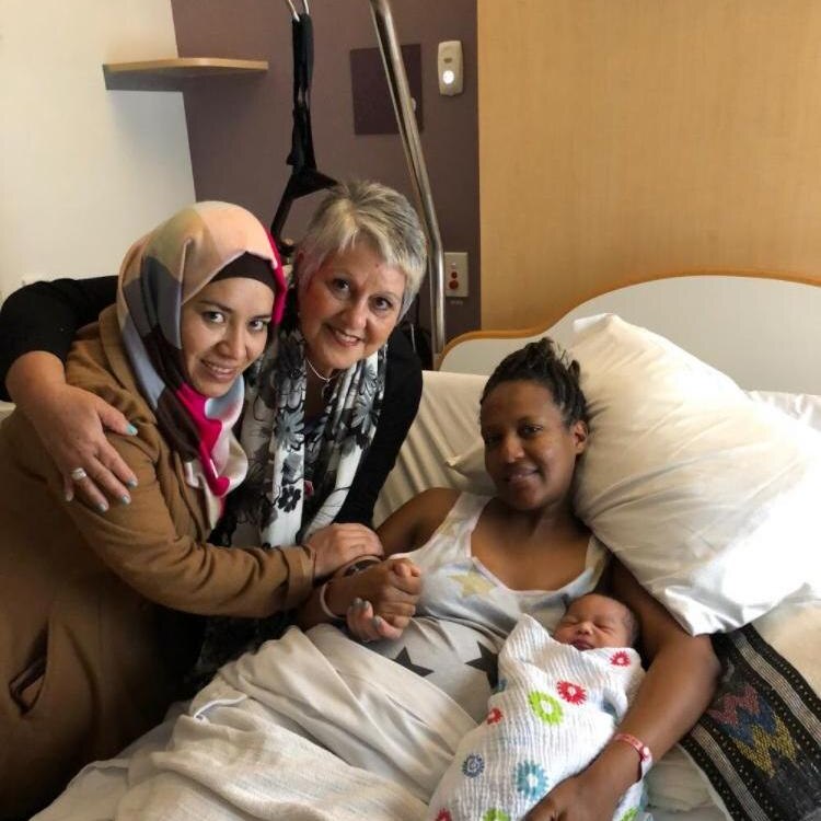 Farzana Muzafari stands next to an older woman, in a hospital room next to a woman in a bed with her baby. All are smiling.