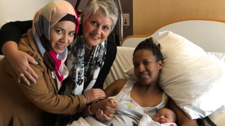 Farzana Muzafari stands next to an older woman, in a hospital room next to a woman in a bed with her baby. All are smiling.
