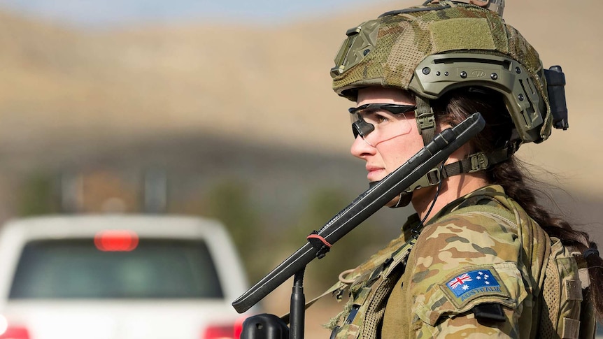 A soldier in combat uniform looks into the distance. She is wearing a helmet and clear glasses