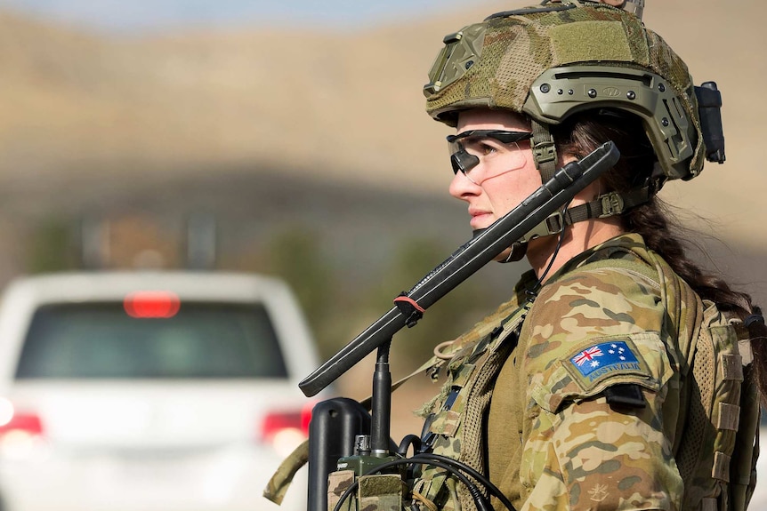 A soldier in combat uniform looks into the distance. She is wearing a helmet and clear glasses