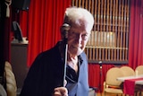 Ron West, an elderly man with white hair, holds up his walking stick while looking at the camera