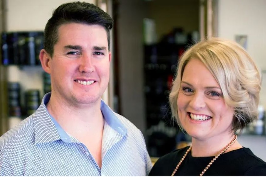 A man and a woman stand side by side smiling at the camera while standing in a salon