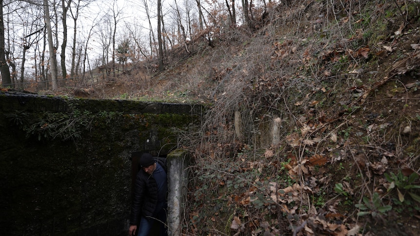 A man walks out of a small built entrance set into the side of a hill covered in leaves and dead plants. 