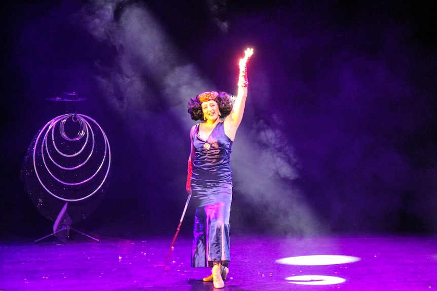 A woman in a purple dress, holds whips under a beam of stage lighting.