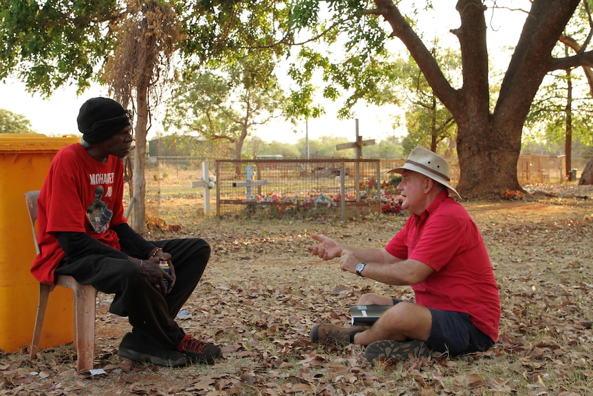 Alan Gray sitting on the ground, talking to an Aboriginal man in a leafy park.