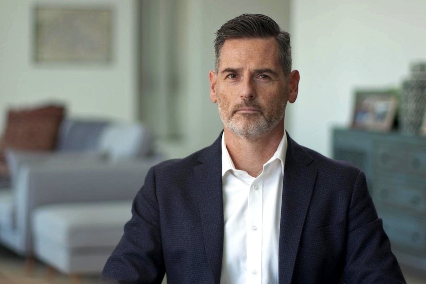 Matthew McQuade sits indoors staring into the camera. He wears a jacket over a white shirt.