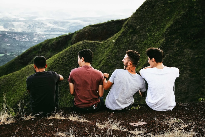 Four males sitting on the ground and laughing against the backdrop of a mountainside