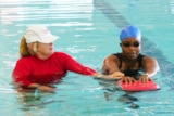 A woman in a red shirt teaching another woman wearing a swimming cap and goggles how to swim. 