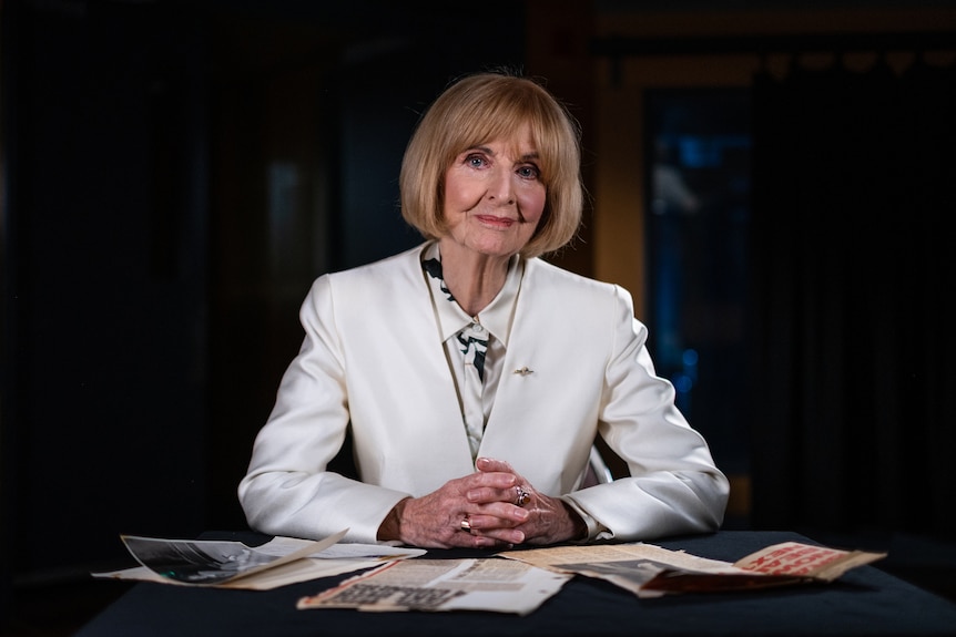 Caroline Jones smiles, her hands clasped in front of her on a table. On the table are old newspaper clippings.