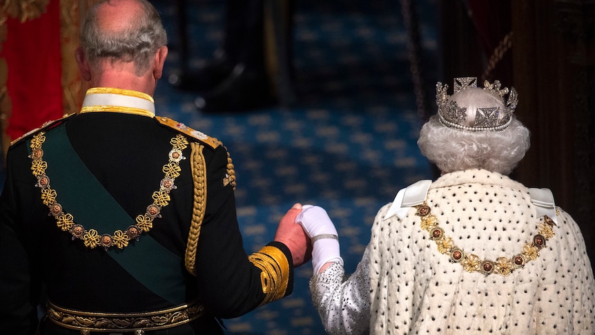 back view, charles holding queen elizabeth's hands, both wearing embellished clothing in hall-like setting 