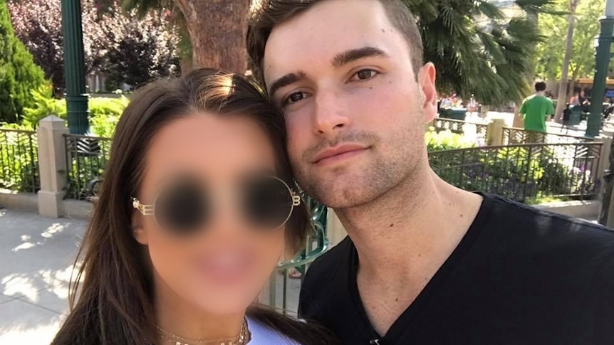 A young man hugs his girlfriend whose face is blurred as they stand in front of a park