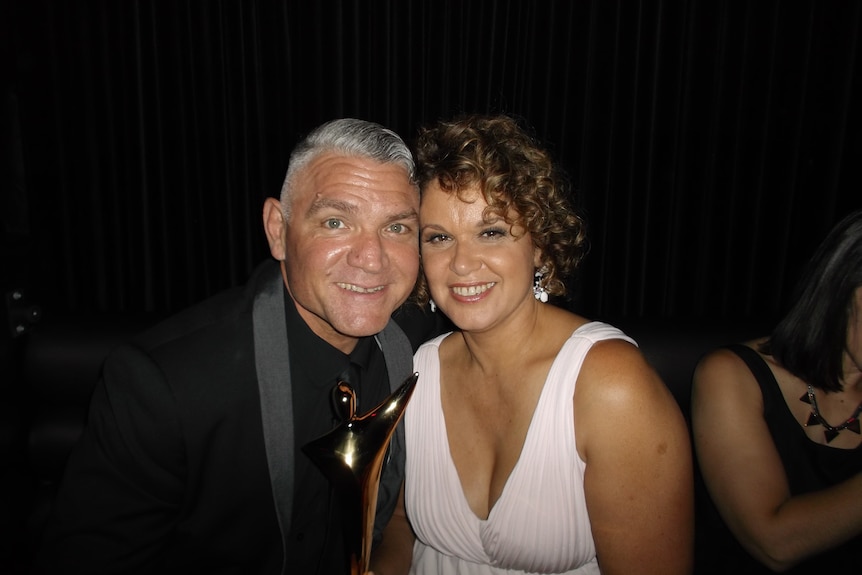 A man with grey hair wearing a suit smiles sitting next to a woman wearing a light blush sleeveless dress