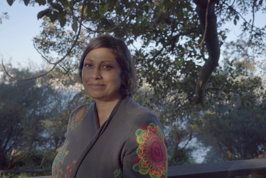 Indira Naidoo has short hair, half smile, wears a grey knit with colourful print, stands under a giant tree. 