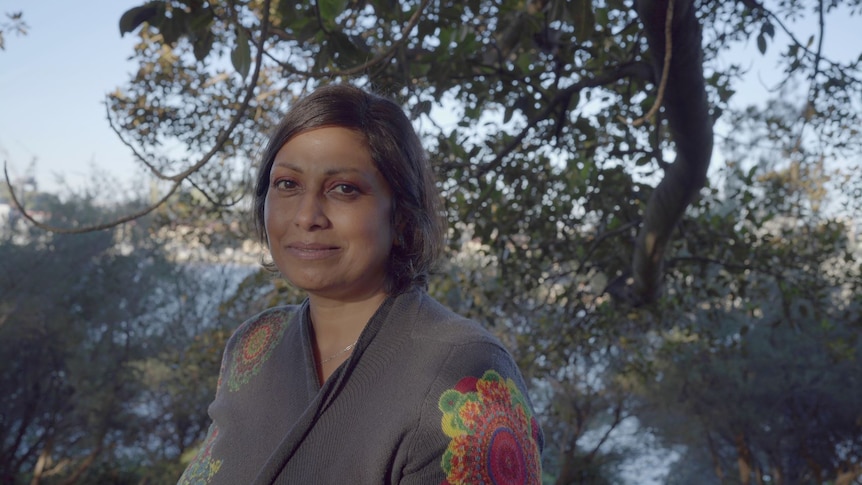 Indira Naidoo has short hair, half smile, wears a grey knit with colourful print, stands under a giant tree. 