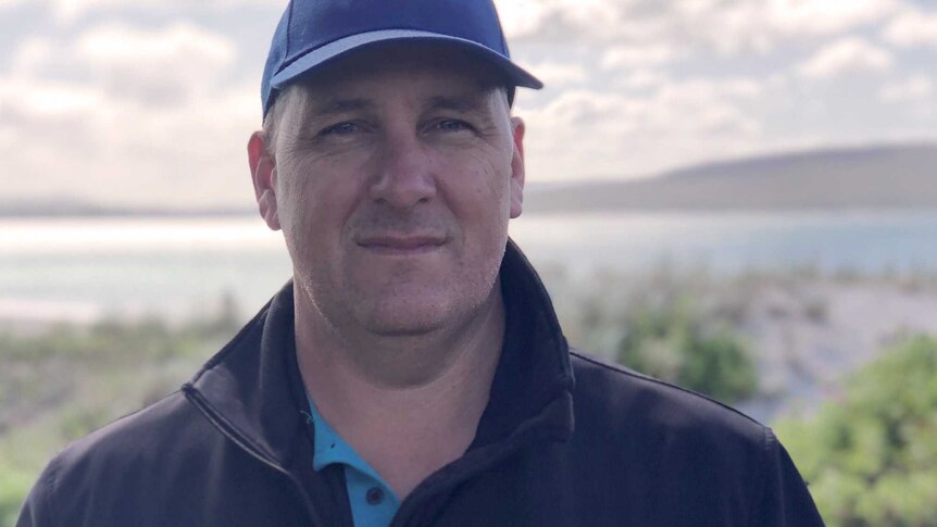 A man in a black jacket and West Coast Eagles cap in a coastal bush location with the sea behind him.