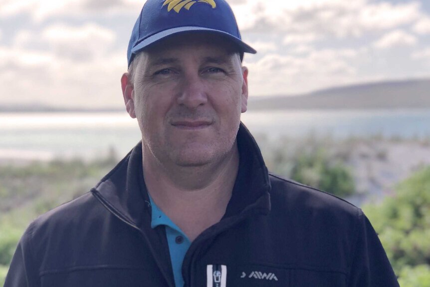 A man in a black jacket and West Coast Eagles cap in a coastal bush location with the sea behind him.