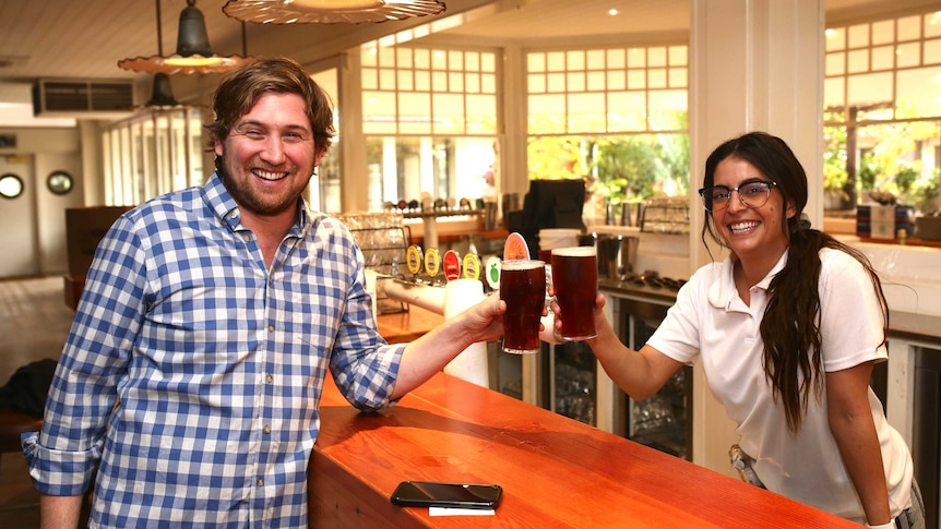 A man and a woman smile at the camera while raising pints of beer at a bar.
