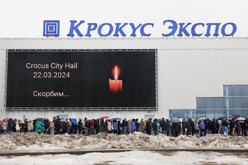 A crowd of people stand in a line beside a building which has a large billboard featuring a candle on the side.