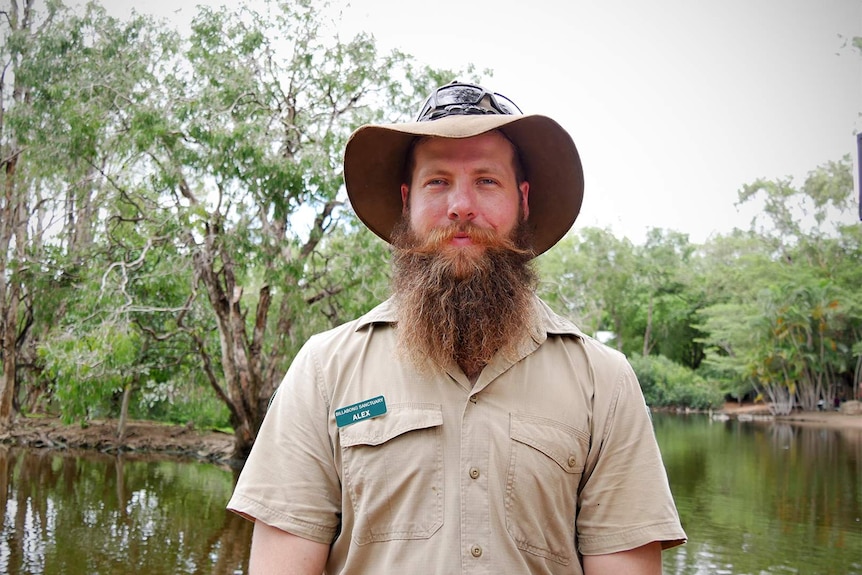 A young man with red facial hair wears a brown ranger uniform and hat in front of a muddy body of water. He smiles for a photo.