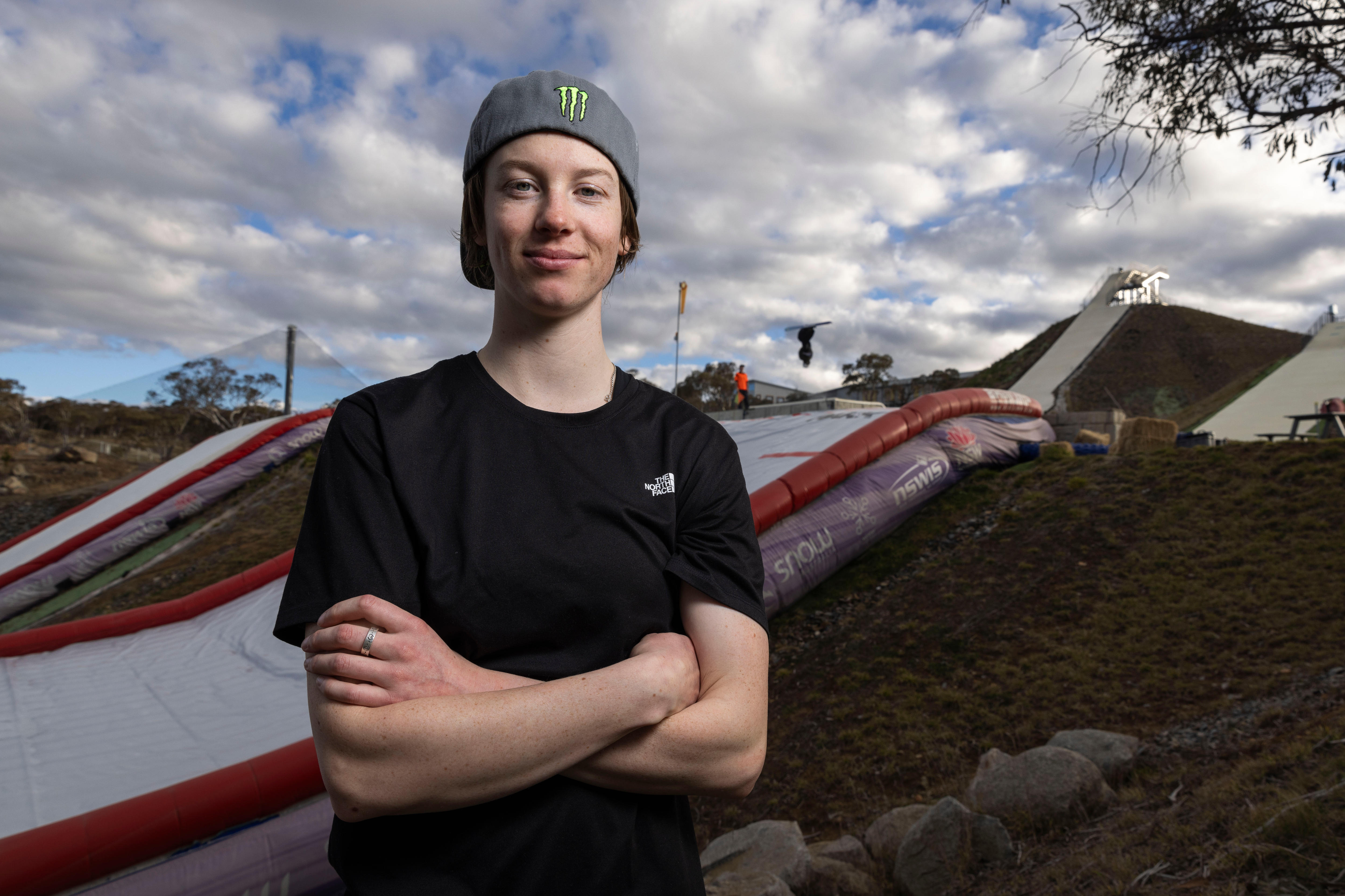 A girl in a beanie and t-shirt, in front of a ski jump. 