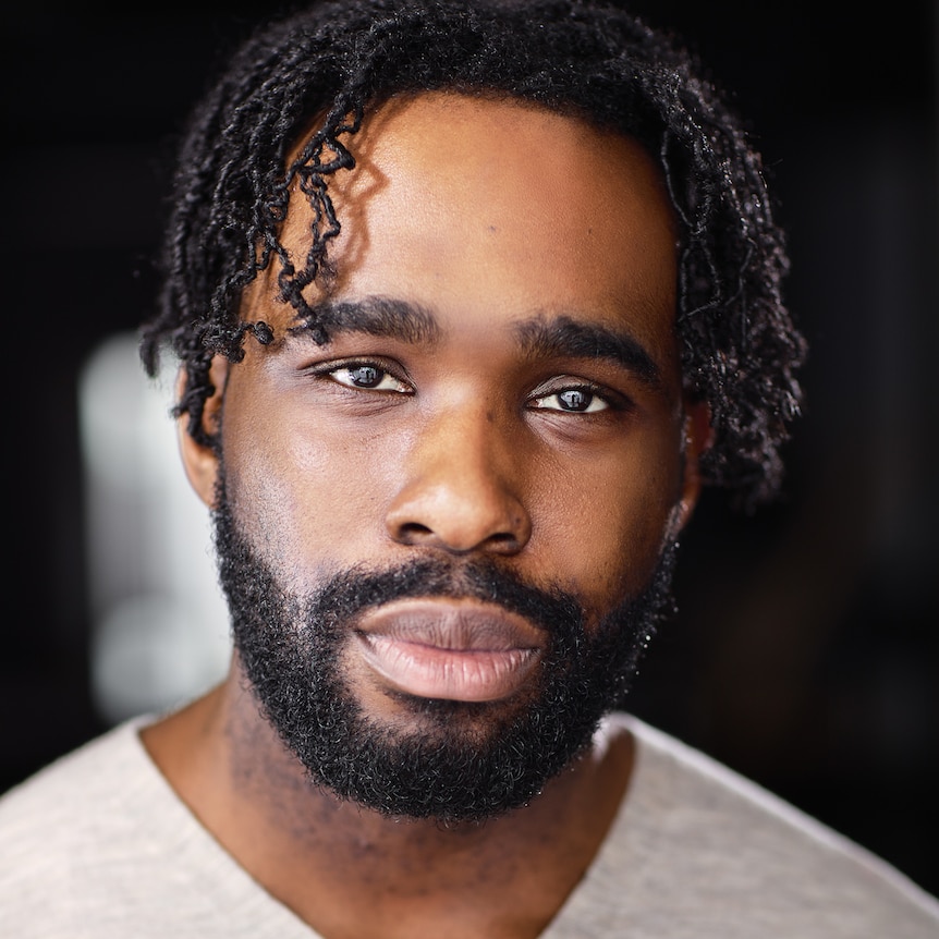 Young man with short black hair and beard and light top, looking at camera..