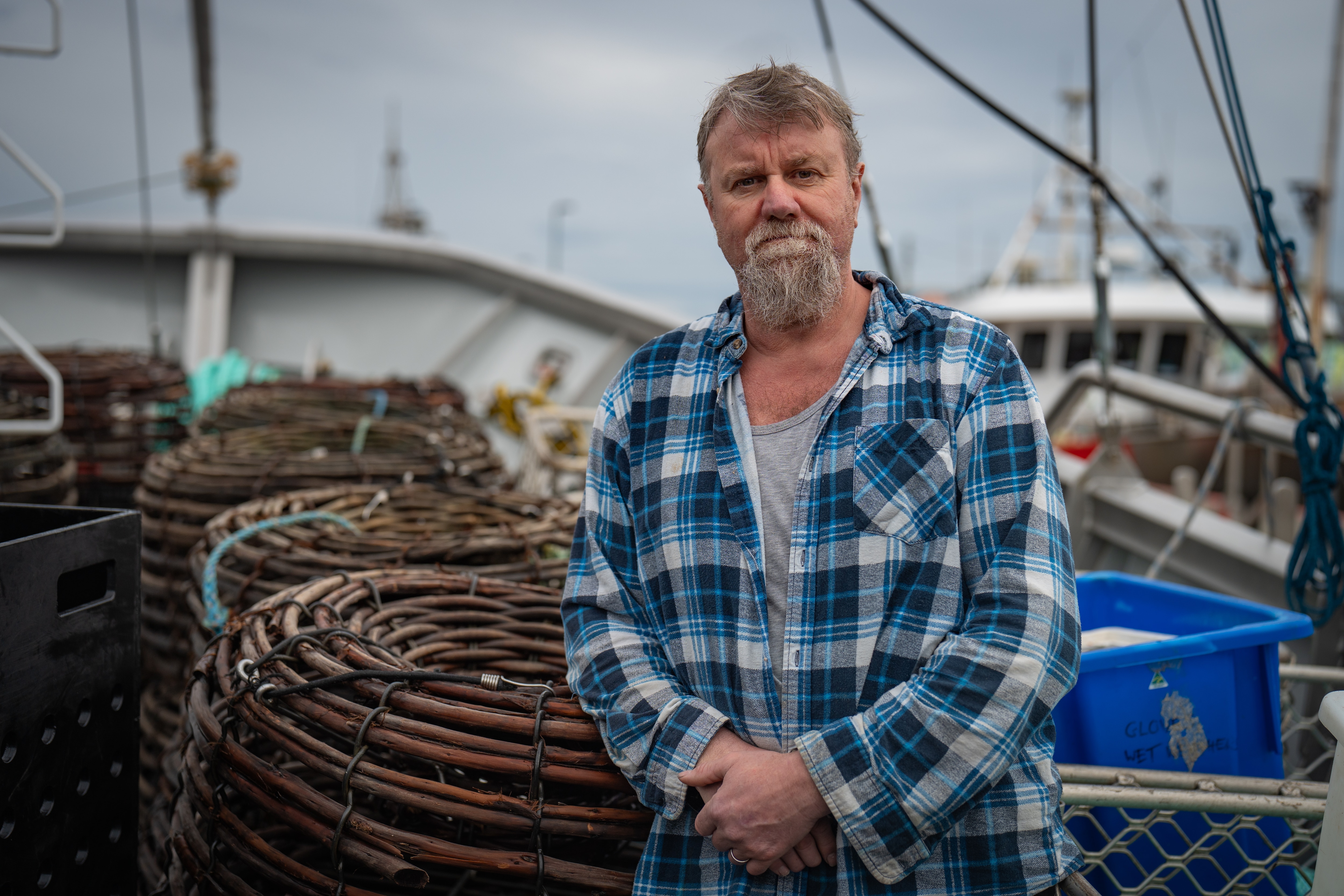 A man in a blue plaid shirt and a beard leans against a stack of lobsters pots