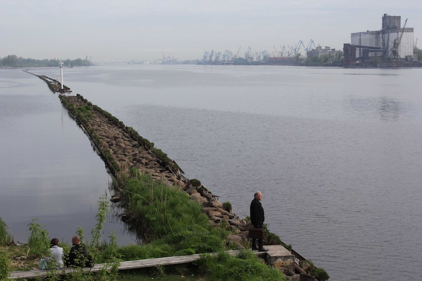 An older man stands alone on a long rocky pier looking out onto a wharf